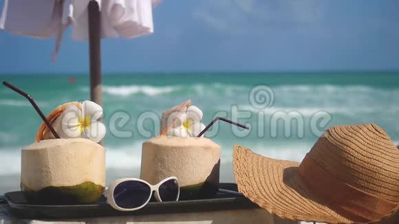 热带新鲜的椰子鸡尾酒在带绿松石海的海滩咖啡馆里装饰着弗兰吉帕尼的花朵和带帽子的太阳镜视频的预览图
