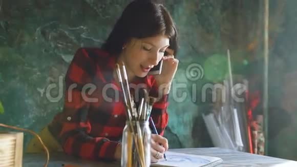 年轻的女艺术家用铅笔在纸笔记本上画女孩微笑打电话视频的预览图