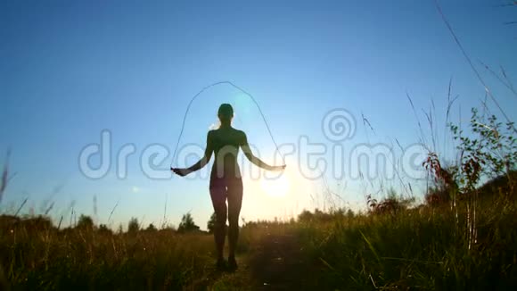 一个美丽的运动的金发碧眼的女孩的轮廓在夕阳下在蓝天的映衬下跳绳视频的预览图