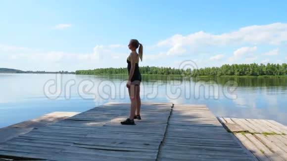 美丽的运动的年轻金发女人伸展做不同的运动跳跃跳跃蹲湖泊河流蓝色视频的预览图