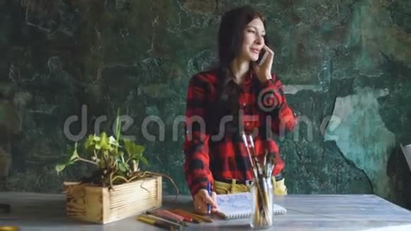 年轻的女艺术家用铅笔在纸笔记本上画女孩微笑打电话视频的预览图
