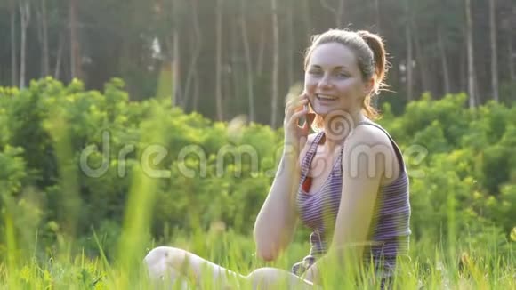 快乐的年轻女子坐在绿色的草坪上在电话或智能手机上交谈视频的预览图