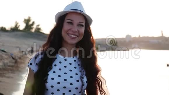 快乐的女孩戴着一顶白色可爱的帽子沿着海边散步在沙滩上看着夕阳微笑着视频的预览图