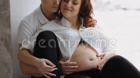 等待孩子出生的年轻夫妇坐在窗边他的手抚摸着她的肚子慢动作视频的预览图