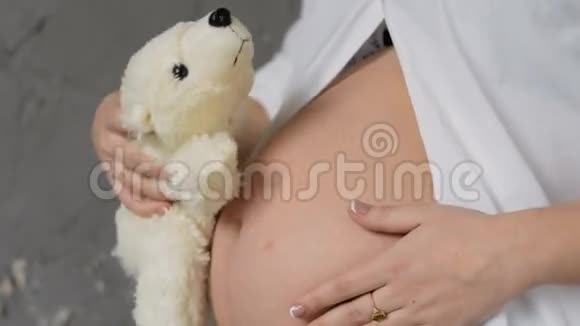 穿白色衬衫的快乐孕妇把泰迪熊放在肚子里泰迪熊听着腹部的声音视频的预览图