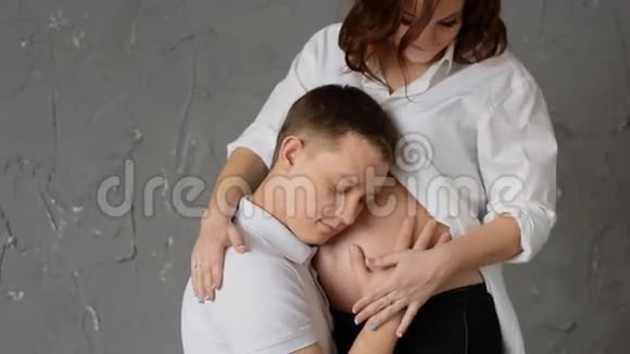 幸福的年轻人听着他怀孕妻子的肚子妻子拥抱丈夫看着镜头视频的预览图