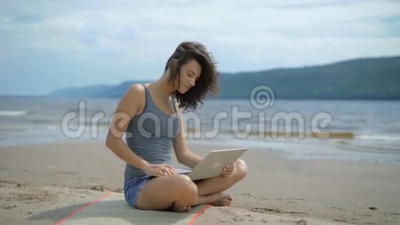 一位年轻迷人的年轻女士正在用笔记本电脑坐在海滩上视频的预览图