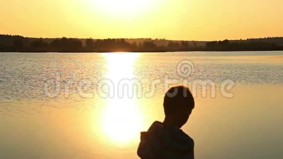 日落时分男孩把石头扔进河里剪影阳光照射在水面上视频的预览图