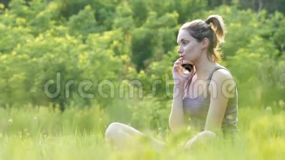 快乐的年轻女子坐在绿色的草坪上在电话或智能手机上交谈视频的预览图