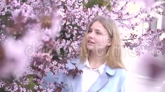 美丽的金发女孩在开满粉红色花朵的树枝上摆姿势的肖像春季视频的预览图