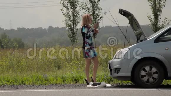 一个金发女孩在乡间小路上的一辆敞篷汽车旁惊慌失措地打着手机视频的预览图