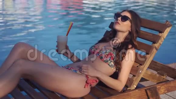 美丽的年轻女子穿着比基尼喝鸡尾酒坐在游泳池旁晒日光浴视频的预览图