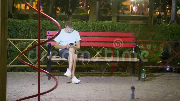 有魅力的悲伤男人坐在一张带智能手机的长凳上在温暖的夏夜在公园里4kStealam射击视频的预览图