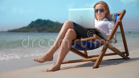 女商人在一个白色沙滩上坐在海边的休息室里用笔记本电脑工作自由职业者或工作狂视频的预览图