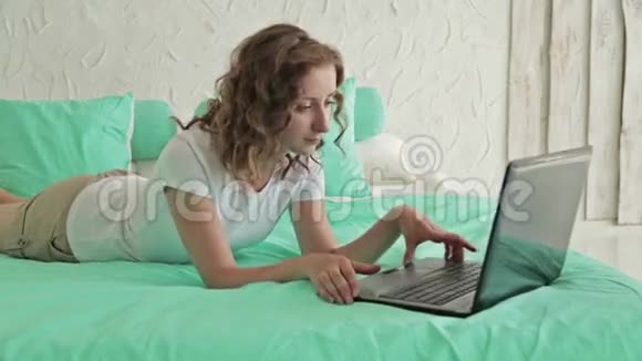 蜷缩的女人躺在床上用笔记本电脑视频的预览图