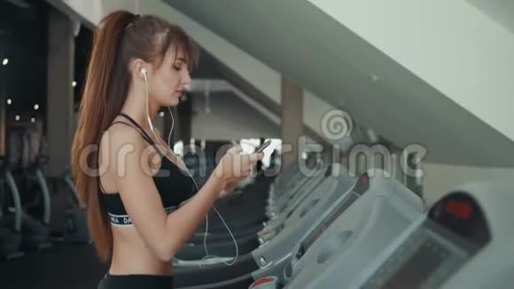 年轻漂亮的女运动员在健身房跑步机上运行智能手机女女孩听手机短信视频的预览图