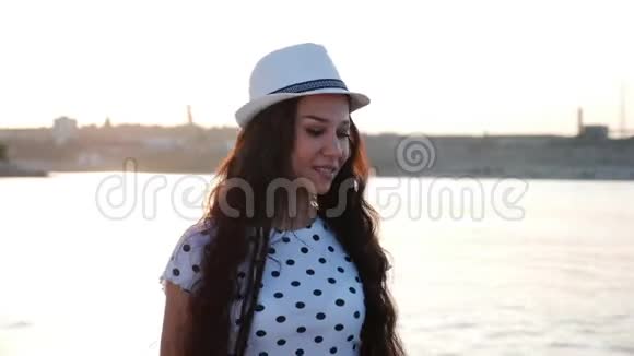 快乐的女孩戴着一顶白色可爱的帽子沿着海边散步在沙滩上看着夕阳微笑着视频的预览图