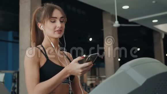 年轻漂亮的女运动员在健身房跑步机上运行智能手机女女孩听手机短信视频的预览图