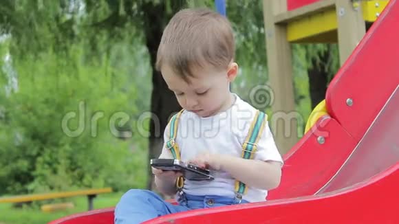 男孩坐在红山上操场上玩电话智能手机视频的预览图