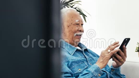 戴着白胡子的亚洲老人在家里用智能手机或平板电脑视频的预览图
