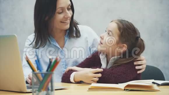 年轻的母女坐在灰色背景的桌子旁在这期间一台灰色笔记本电脑正在观看母亲视频的预览图