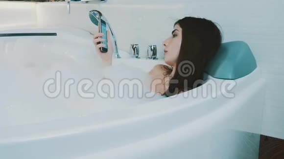 布鲁内特女孩在充满泡沫的浴缸里放松打开智能手机享受休息视频的预览图