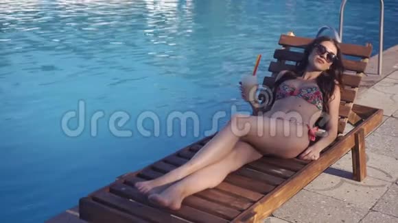 美丽的年轻女子穿着比基尼喝鸡尾酒坐在游泳池旁晒日光浴视频的预览图