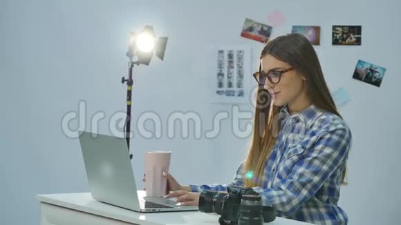 年轻漂亮的女摄影师在办公室里用手提电脑和喝咖啡视频的预览图