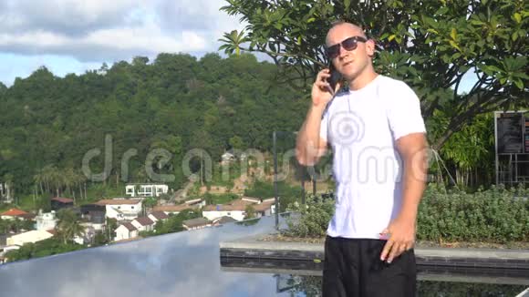 站在屋顶上的无限游泳池里一位英俊的年轻人在手机上交谈时的户外画像视频的预览图