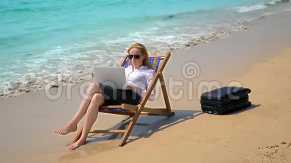 女商人在一个白色沙滩上坐在海边的休息室里用笔记本电脑工作自由职业者或工作狂视频的预览图