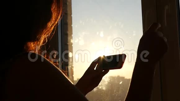日落时分年轻漂亮的女孩在靠近窗户的地方开着手机动作缓慢视频的预览图