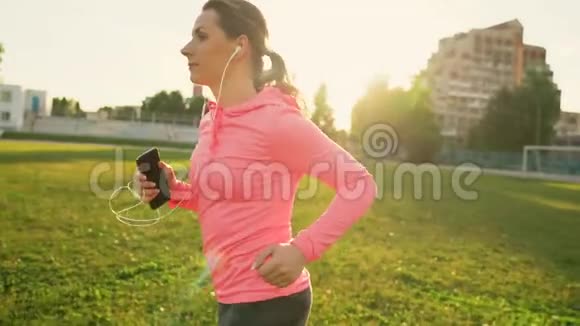 日落时分戴着耳机和智能手机的女人穿过体育场慢动作视频的预览图