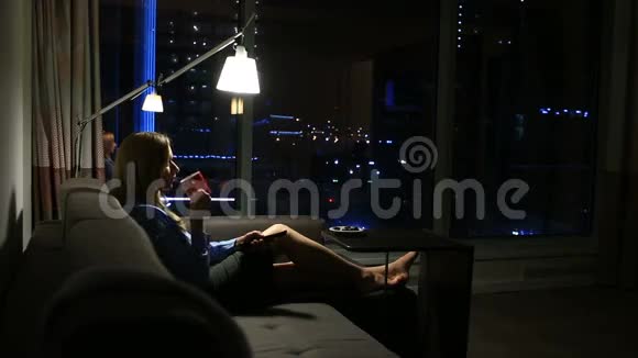 一个女人躺在沙发上手里拿着一个杯子靠近窗户可以看到城市的夜景看电视视频的预览图