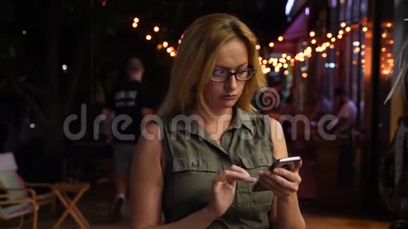 夏天一个金发女人在黑暗中在智能手机上键入一条短信晚上晚上4k慢动作视频的预览图
