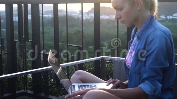 一个漂亮的金发女人坐在电脑上享受清晨在屋顶上叽叽喳喳地唱歌视频的预览图