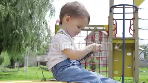 一个小男孩坐在长凳上在操场上玩智能手机微笑着视频的预览图