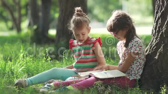 孩子们坐在树旁的草地上看一本书两个小女孩一起玩视频的预览图