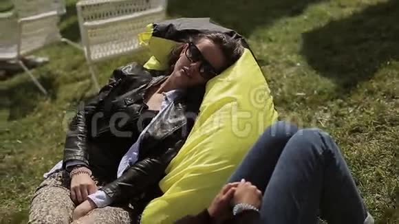 两个戴太阳镜的女孩在绿草上的黄色豆袋上放松微笑夏季节日休息视频的预览图