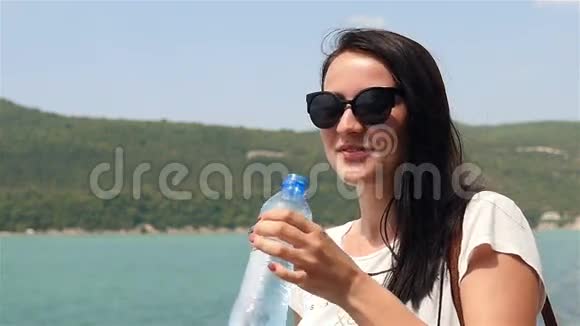 阳光明媚的夏日年轻女子在山湖的背景下喝着纯净的水视频的预览图