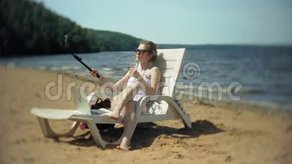 一个穿着白色比基尼的年轻女孩躺在沙滩上的躺椅上晒太阳还在智能手机上拍照视频的预览图