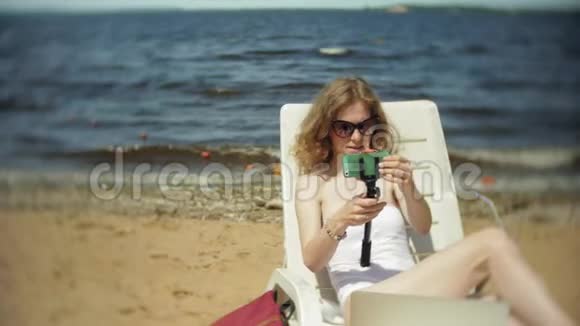 一个穿着白色比基尼的年轻女孩躺在沙滩上的躺椅上晒太阳还在智能手机上拍照视频的预览图