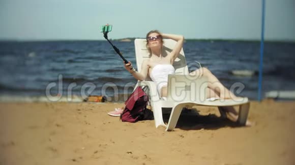 一个穿着白色比基尼的年轻女孩躺在沙滩上的躺椅上晒太阳还在智能手机上拍照视频的预览图