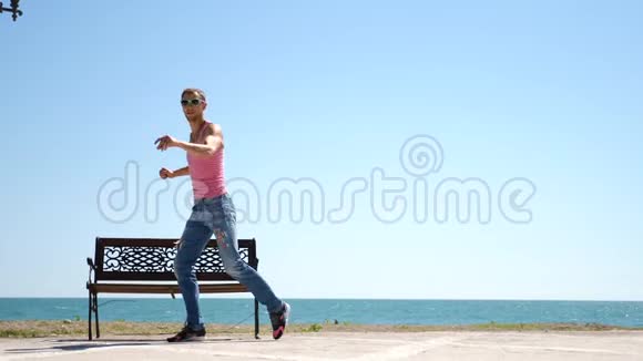 一个年轻快乐的家伙跳着现代芭蕾在海边的背景下摇摇晃晃地走着缓行视频的预览图