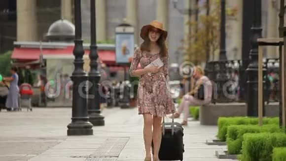 快乐的年轻女游客拿着票和手提箱走在城市的街道上视频的预览图