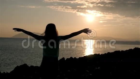 美丽的年轻女性在夕阳下仰望大海和举起双手的剪影视频的预览图