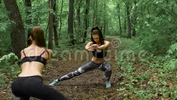 林中两个身材修长的健身少女布鲁内特在路径上练习他们执行蹲来伸展视频的预览图