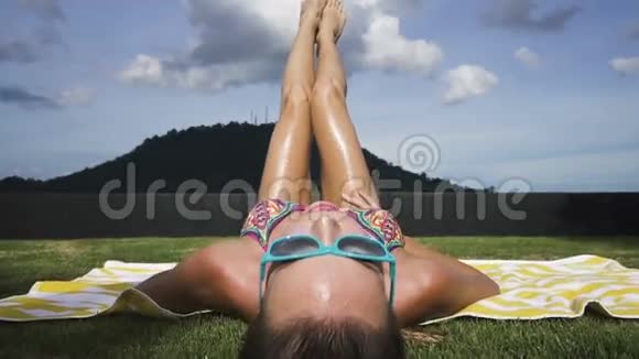 年轻的女人穿着比基尼和蓝色太阳镜躺在条纹海滩毛巾上腿在绿色草地屋顶的空气中视频的预览图