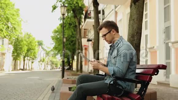 戴眼镜坐在长凳上户外使用平板电脑的漂亮时髦男人视频的预览图