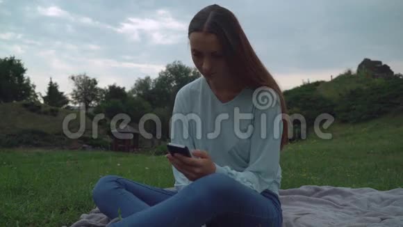 一个年轻的女孩坐在公园草坪上的毯子上在电话里得到了一条信息视频的预览图