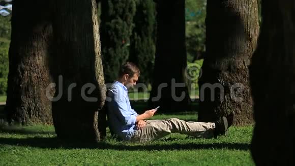 帅哥坐在棕榈树下用智能手机上网视频的预览图